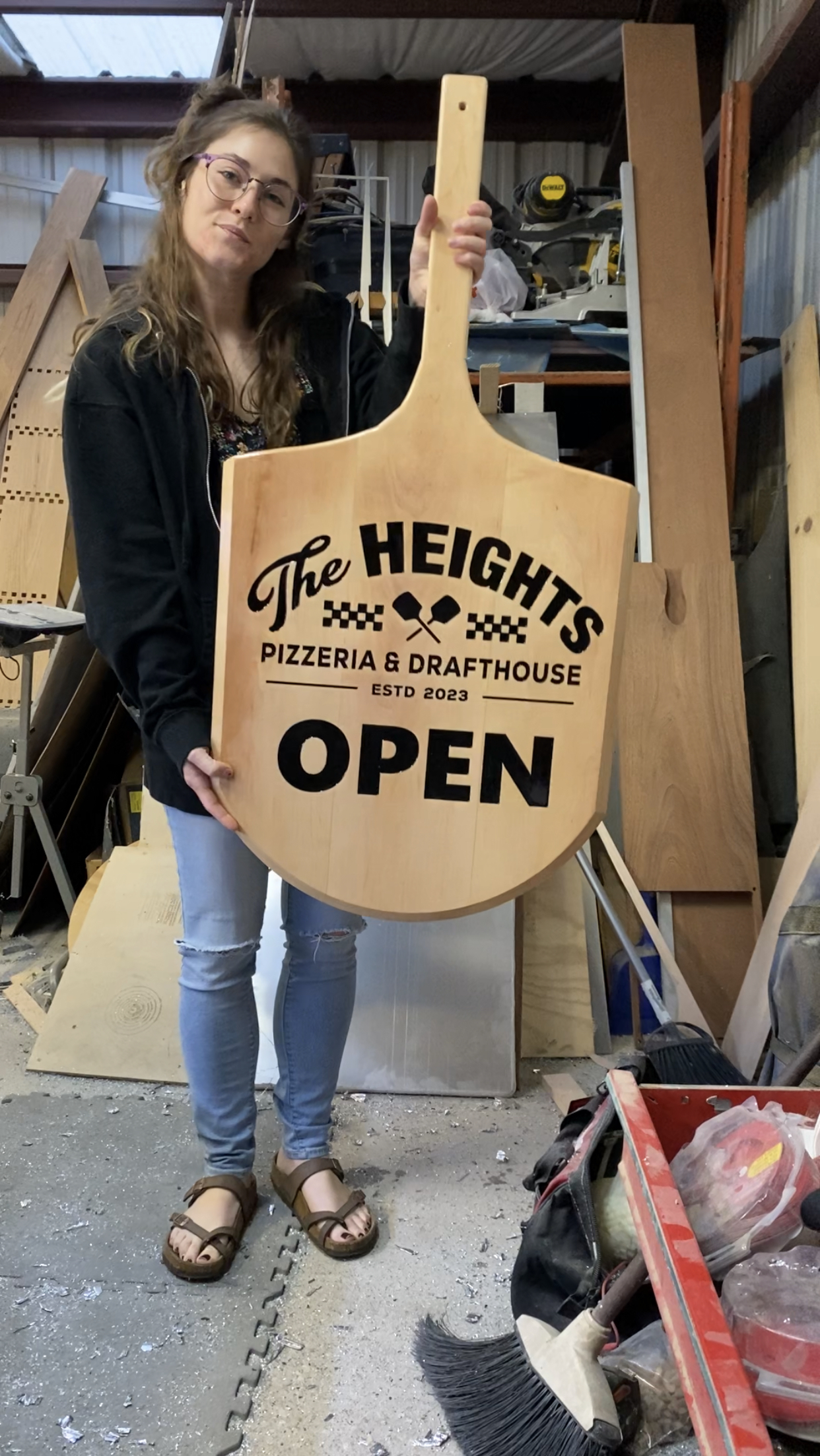 woman holding a pizza paddle shaped wooden sign that says "The heights pizzeria & draughthouse OPEN"