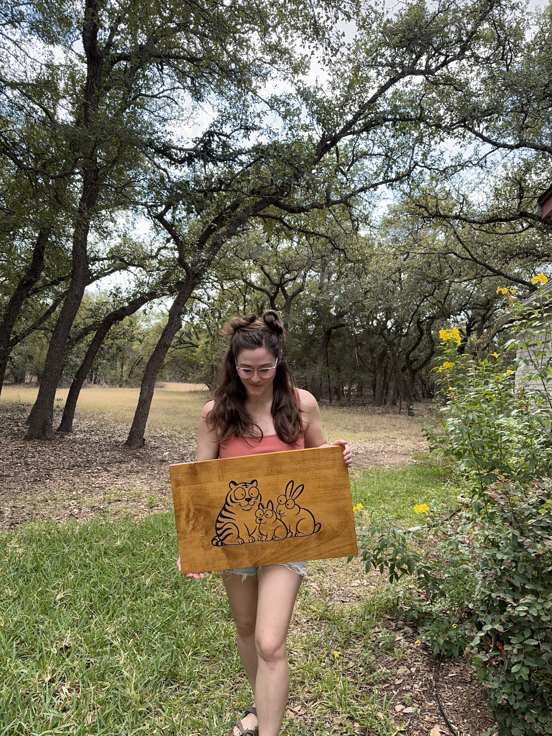 woman holding a wooden sign with a carved image of an animal family of 3, trees and flowers in the background